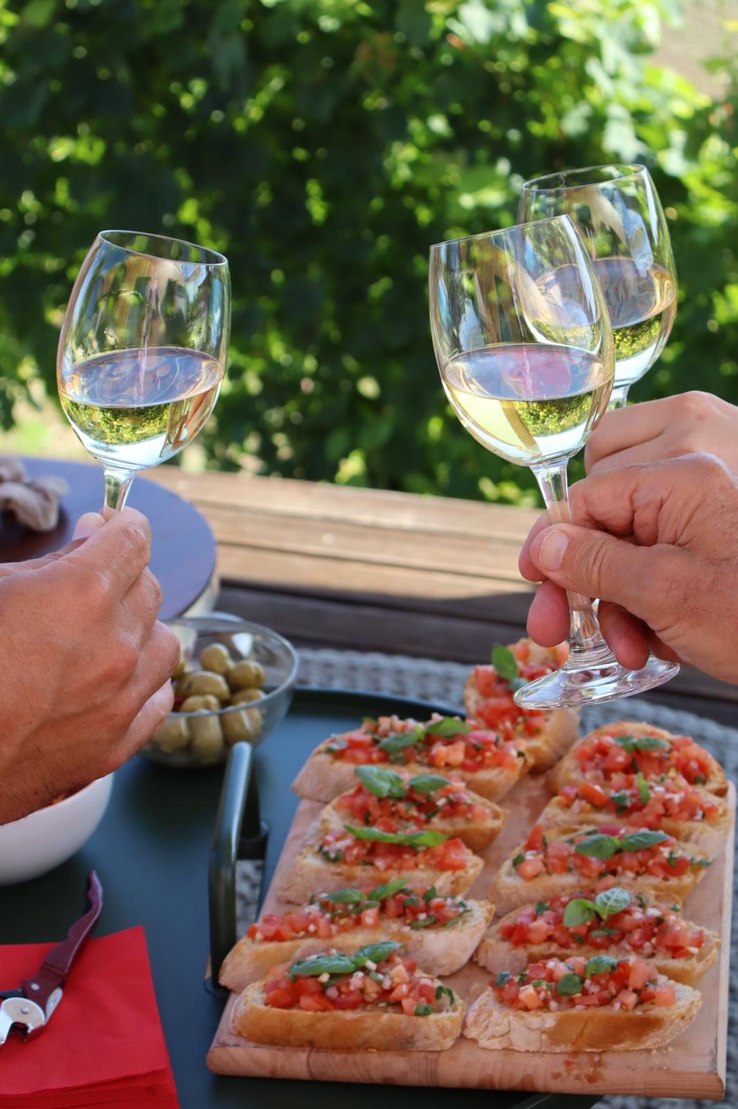 Anstossen und Apero Drei gefüllte Weissweingläser stossen an. Im Hintergrund sieht man eine Bruschetta